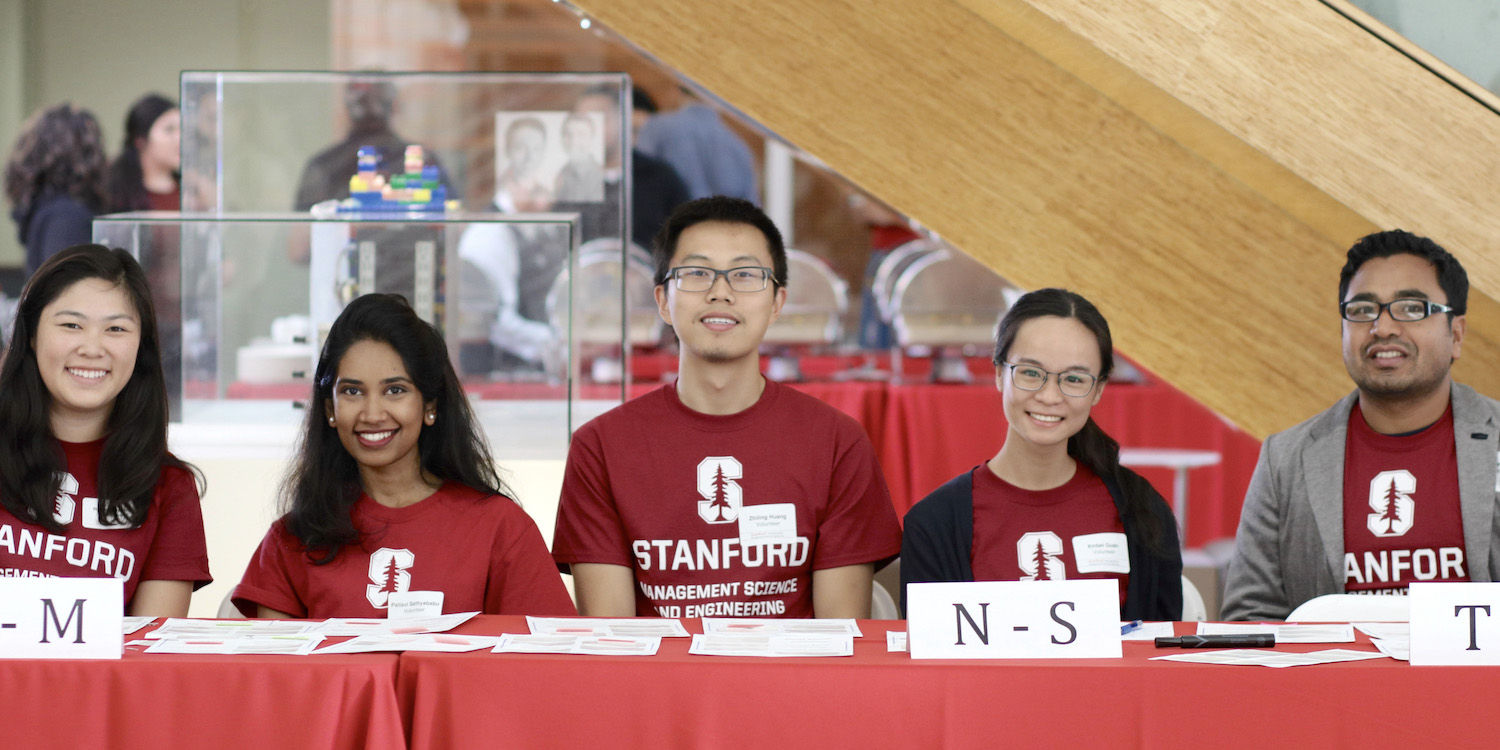 five students in Stanford t-shirts sit behind a table with welcoming expressions